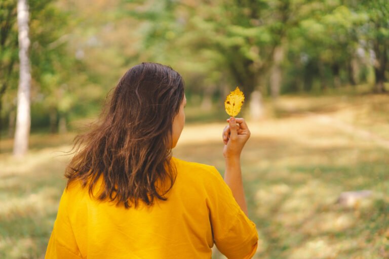 A woman enjoying a sunny autumn day while holding a yellow leaf in a peaceful park
